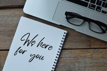 Top view of eyeglasses, laptop and notebook written with text WE HERE FOR YOU over wooden background. 
