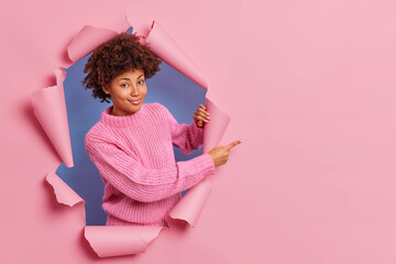 Pleased dark skinned young woman with Afro hair wears casual sweater peeping through paper background indicates away on copy space for your promotion or advertisement. Look at this good offer