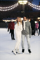 Happy stylish couple of lovers having fun, looking away, standing at ice skating rink outdoor. St. Valentines Day, Winter leisure, New Year holidays concept.