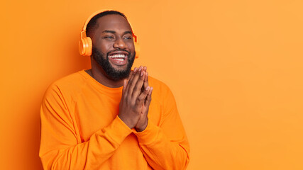 Happy plump dark skinned man keeps palms pressed together smiles broadly dressed in casual sweater wears stereo headphones listens pleasant melody poses against orange studio wall with copy space