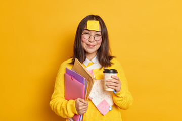 Positive Asian female worker holds disposable cup of coffee holds folders has sticker with graphic...