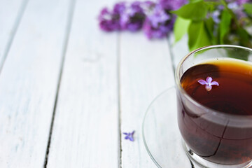 Delicate setting of the morning tea table with lilac flowers, a transparent cup of tea and a saucer and a white vase on a white wooden board. Copy space. Spring breakfast concept