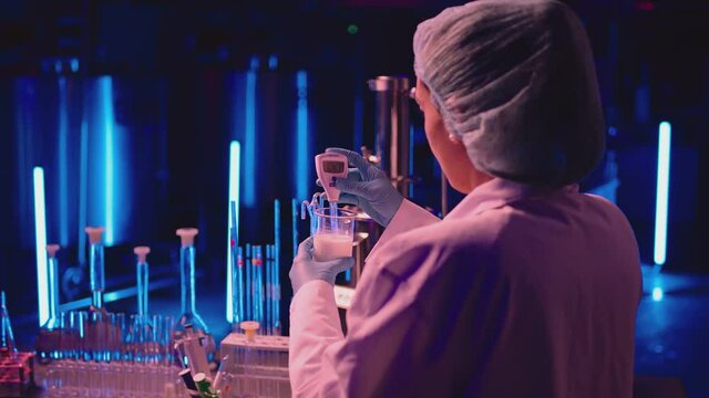 Woman dressed on white lab coat, working on laboratory with test tubes on blue light background. Worker testing the quality of milk by digital multimeter . Milk production approval process in factory