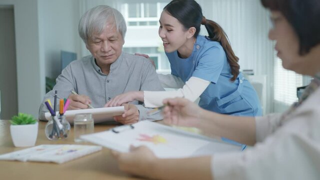 Attractive young senior asian citizen couple sit at home watercolor painting artwork on desk table with nurse feeling happy in mental health therapy or asia older people quarantine activity lifestyle.