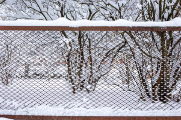 Snow on a steel mesh fence of red color against a background of trees