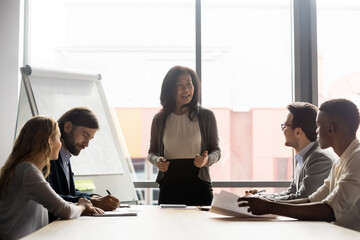 Happy mature korean asian female leader holding negotiations briefing meeting with motivated employees in modern office. Smiling skilled mixed race businesswoman mentoring colleagues near flipchart.