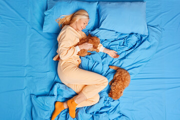 Pleased mature woman stays in bed plays with puppies loves animals dressed in domestic clothes...
