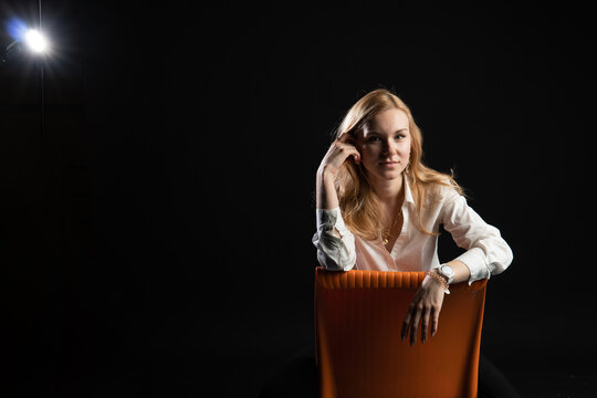 teenager sits on an orange chair. with a charismatic appearance, on a black background in the studio, in blazer business woman, blonde