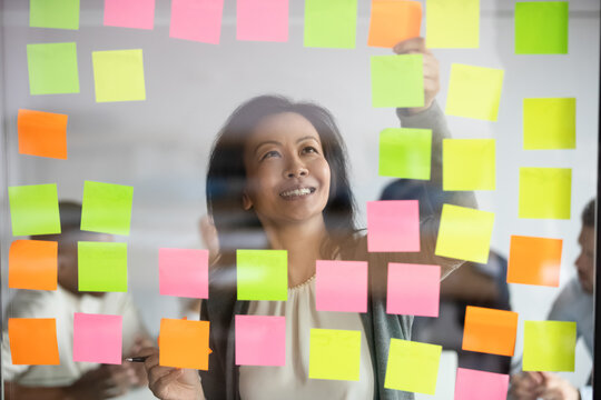 Happy middle aged korean female project manager standing behind glass wall, adding colored sticky notes on window, organizing working processes or adding steps, developing corporate strategy.