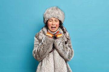 Sad frustrated young Asian woman wears fur hat and coat cries unhappily dresses for cold weather expresses negative emotions isolated over blue backgrund. Northern inhabitant adapted to frost