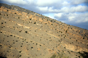View of the mountain wall with little vegetation on the many rock layers and cloudy blue sky in the background, Jabal Akhdar, towards Nizwa, Oman