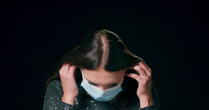 Close-up of a teenage caucasian girl wear brocade dress putting on a protective medical face mask against a black studio background with copy space.