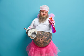 Bearded surprised obese man with tattooed belly holds detergent and plunger does household duties...