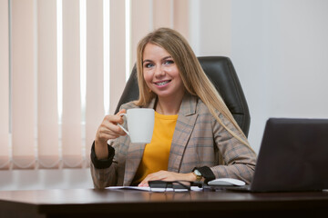 Young cheerful lady boss is holding a cup of coffee in her office. Coffee break