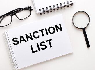 SANCTION LIST is written in a white notebook on a light background near the notebook, black-framed glasses and a magnifying glass.