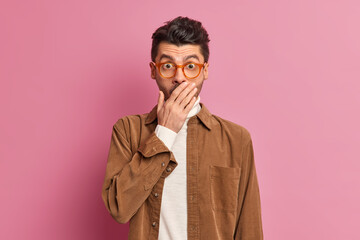 Obraz premium Waist up shot of impressed unshaven guy covers mouth stares at camera feels very surprised wears spectacles and brown shirt poses against pink background. People human reaction and emotions concept