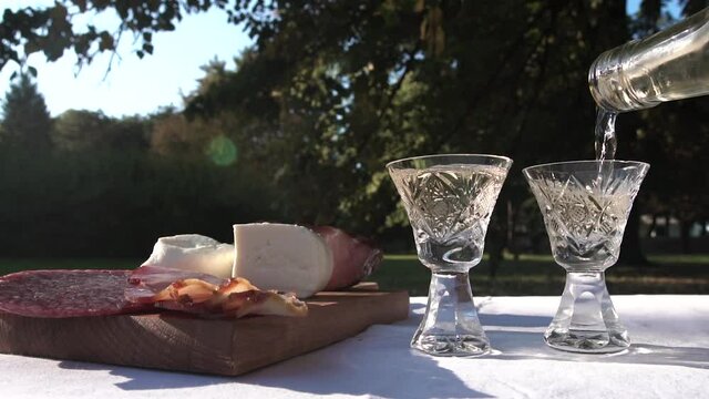 Pouring Rakija Brandy in Small Glasses on Picnic Table With Cheese and Sausage