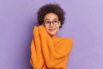 Lovely ethnic girl with natural Afro hair keeps hands near face and looks with dreamy expression somewhere wears loose orange knitted sweater isolated over purple background. Pleasant feelings