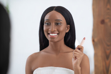 Smiling millennial african american lady with perfect skin looks in mirror and holds nude lipstick