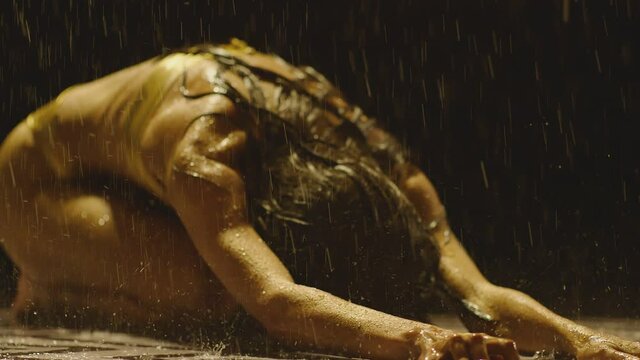 Sexy and beautiful brunette girl on bikini kneeling with emotional movements on the wet tiles under the heavy rain in bathroom interior in slow motion . Show of sensual movement inside rainy room .