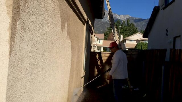 A cleaning service or painter tradesman pressure washing a concrete wall, starting from the bottom and working his way up.