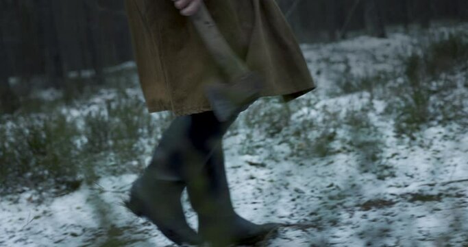 Scary man with a cape and an olde axe in hands walking in woods in winter. Scary horror movie concept. 4k slow motion.