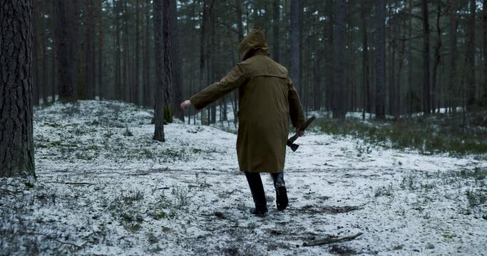 Scary man with a cape and an olde axe in hands walking in woods in winter. Scary horror movie concept. 4k slow motion.