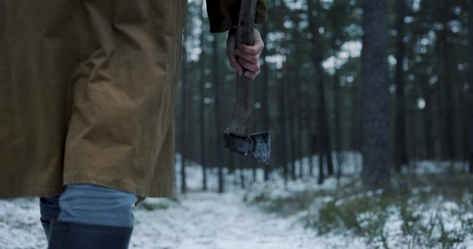 Scary man with a cape and an olde axe in hands walking in woods in winter. Scary horror movie concept. 4k slow motion.