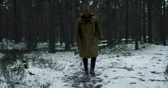 Scary man with a cape and an olde axe in hands walking in woods in winter. Scary horror movie concept. 4k slow motion.