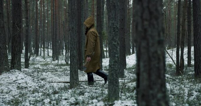 Scary man with a cape and an olde axe in hands walking in woods in winter. Scary horror movie concept. 4k slow motion.