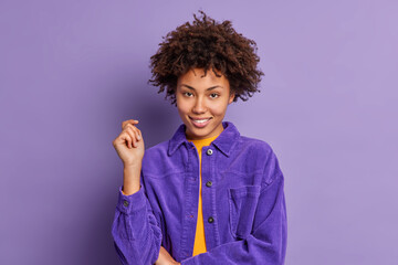 Pretty self confident female model with Afro hair keeps hand raised smiles gently looks directly at camera listens attentively interlocutor wears stylish velvet jacket isolated over purple background