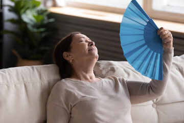 Close up exhausted middle aged woman waving blue paper fan, leaning back on couch at home,...