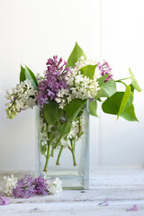 A bouquet of spring purple and white lilacs in a vase on a light background of wood and tiles. Glass transparent container with water. Aromatic composition. Soft focus