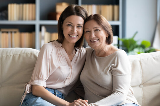 Head shot portrait mature woman with grown up daughter hugging, holding hands, sitting on cozy couch at home, smiling young female with older mother posing for family photo, enjoying leisure time