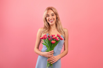 Portrait of romantic blonde woman in dress smiling and holding spring tulips on pink background. Holiday celebration