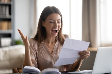Close up overjoyed young woman reading good news in letter, holding paper sheet, sitting at desk...