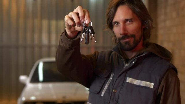 Handsome bearded man showing car keys and teasing in garage with white car in blurred background