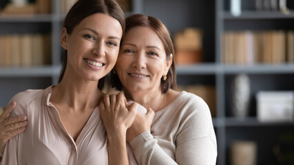 Head shot portrait happy two generations of women hugging, standing at home, smiling mature elderly...