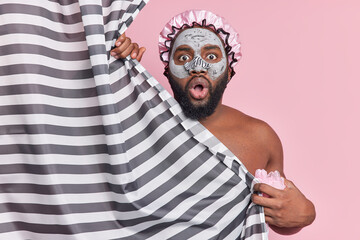 Indoor shot of surprised bearded man takes shower in bathroom wears water proof bath hat hides...