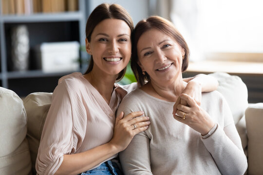 Head shot portrait smiling young woman hugging mature mother, sitting on couch together, happy middle aged mum and adult grownup daughter looking at camera, posing for family photo at home