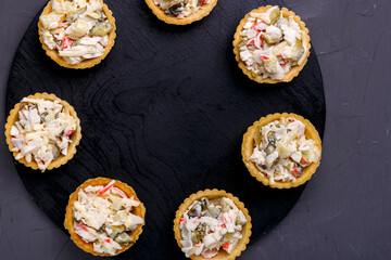 Tartlets with salad with pineapple and shrimps on a black wooden round board on a gray concrete background copy space
