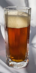 Semi-dark beer and foam in a faceted glass glass close-up, illuminated by the sun on a white background