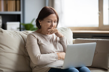 Close up thoughtful mature woman looking at computer screen, touching chin, sitting on couch with laptop on laps, reading bad news in email, thinking about finance or health problem, having doubts