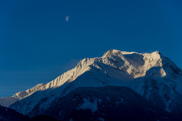 Dark mountain with sunlit snowy top - the moon is visable in early afternoon