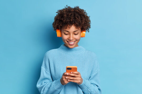 Smiling teenager with Afro hair listens favorite music track holds mobile phone downloads song to her playlist dressed in casual jumper isolated over blue background. People hobby technology