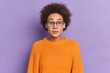 Fototapeta na wymiar Portrait of stunned curly millennial girl stares through spectacles dressed in orange sweater reacts on shocking news poses over vivid purple background. Astonished Afro American female holds breath