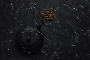 Metal black teapot and dry tea leaves on a black stone background. Asian cast iron teapot. Top view, flat lay. Selective focus