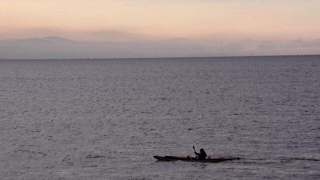 Alone kayaker moves across fjord at sunset in silhouette, long shot