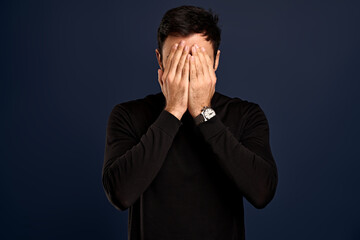 Distressed, tired businessman in black high neck sweater, hiding face, facepalm, holding hands on head and sighing, feeling troubled, exhausted as working hard during holidays, stand Blue background
