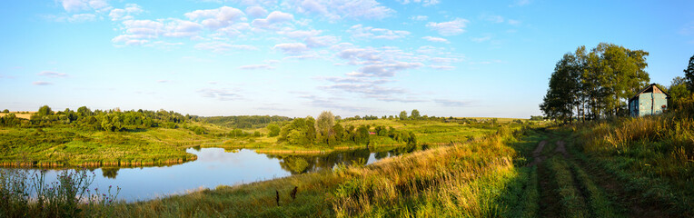 Fototapeta na wymiar Sunny panoramic coyntryside landscape with rural road and calm river during august morning